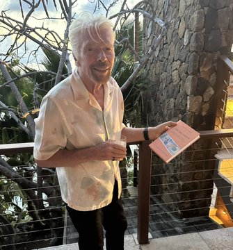 Sir Richard Branson, the billionaire entrepreneur and founder of the Virgin Group, smiles while reading a book titled "The Big Print" by Lawrence Lepard, which focuses on Bitcoin. He's dressed casually in a light-colored patterned shirt and appears to be at an outdoor location with lush greenery and a stone wall in the background. The caption humorously suggests that the wealthy business mogul's interest in the Bitcoin-focused book signals a bullish trend for cryptocurrency adoption among the ultra-wealthy.