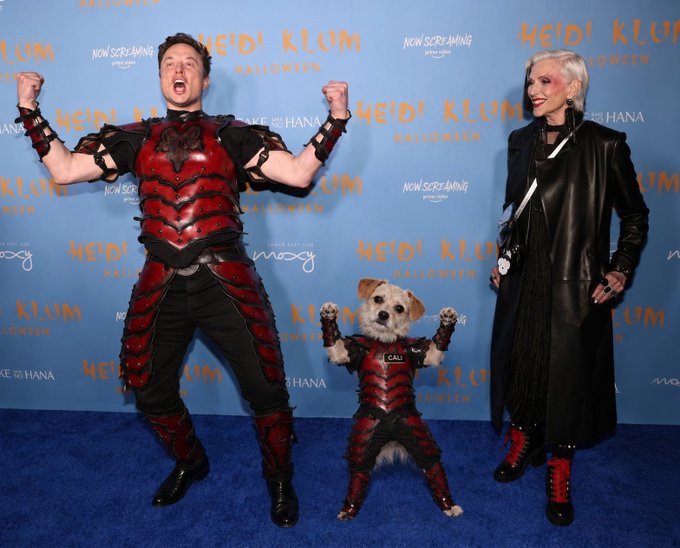 A man in a red and black armored costume and a woman in a black leather outfit pose on a blue carpet at the 'Heidi Klum Halloween' event backdrop, alongside a small dog in a matching red and black armor costume.