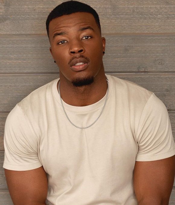 A portrait of a man with short dark hair and a goatee, wearing a beige t-shirt and a silver chain necklace, posed against a wooden plank background.