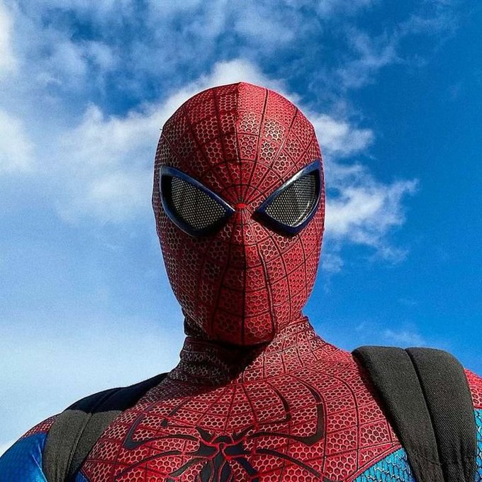 A close-up of a person in a Spider-Man costume with a textured red mask and large eye lenses against a blue cloudy sky.