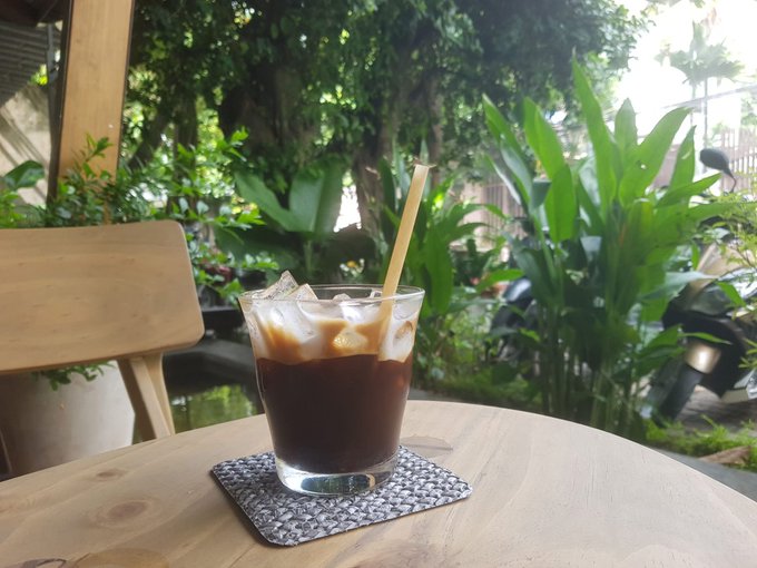 A glass of iced coffee with a bamboo straw sits on a woven coaster atop a wooden table, with a lush garden of tropical plants in the background.