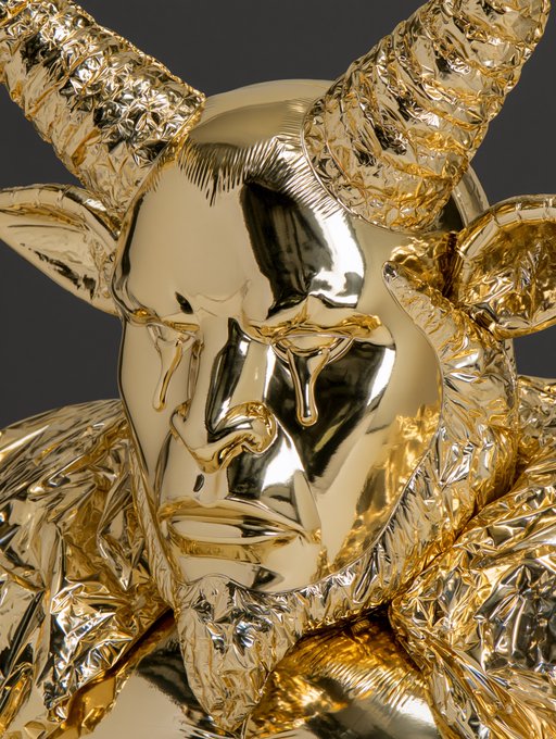 A highly reflective, ornate golden sculpture of a face with horns is shown against a black background.