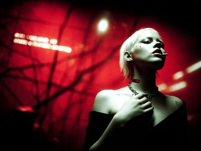 A cinematic, red-lit photograph of a platinum blonde woman with a nose ring, wearing a black off-shoulder top and a necklace, with visible tattoos on her collarbone and shoulder.