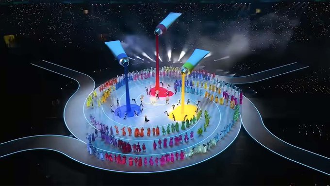 The image features a large group of people dressed in colorful costumes, standing around a circular platform with blue and red lights. The crowd appears to be part of an event or performance, possibly related to the Olympics. There are at least 13 visible individuals in the scene, all wearing vibrant outfits that contrast against the dark background.

The image also includes text elements such as "Never forget the 2 blue and red (Purple) Proton cauldrons at the 2026 Olympics Also the Quarks floor design $XPR". This suggests that there might be a connection between the colorful costumes of the people in the image and some sort of event or performance related to the Olympics. The presence of these elements indicates that this scene is likely part of an advertisement, promotional material, or a visual representation of a specific aspect of the Olympic Games.