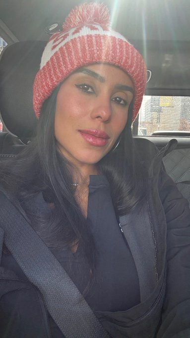 A selfie of a woman with long dark hair wearing a red and white knit pom-pom beanie, sitting in a car with a seatbelt across her chest.