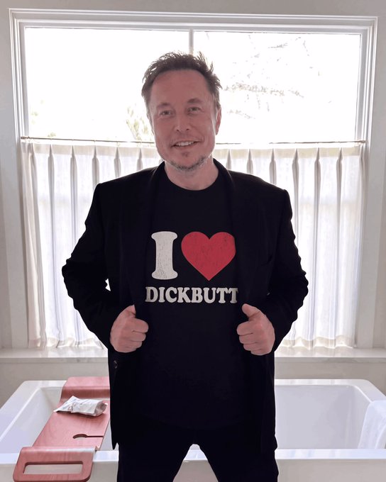 A man standing in front of a window wearing a black blazer and a black t-shirt with 'I ♥ DICKBUTT' printed on it.