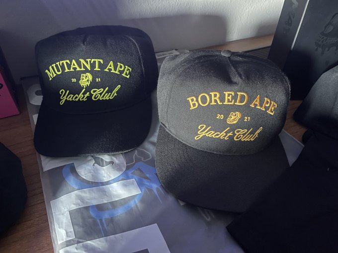 Two baseball caps, one black labeled 'MUTANT APE Yacht Club 2021' and one gray labeled 'BORED APE Yacht Club 2021', sit on a surface with graphics.