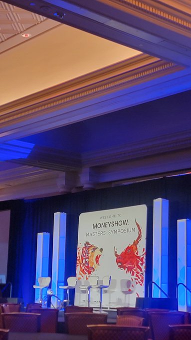 The image shows the MoneyShow Masters Symposium stage in Las Vegas, featuring illuminated pillars and a banner with the text "WELCOME TO MONEYSHOW. MASTERS SYMPOSIUM" decorated with fiery bull and bear graphics. The empty stage with bar stools set up for panelists relates to the post's mention of preparing to talk crypto and investing at the MoneyShow event.
