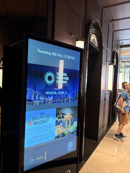 A digital signage display in a hallway showing 'Tuesday, 06 Oct, 12:38 PM', an advertisement for the 'Global Onchain Summit' at Oculus, Level 3, and a promotion for 'The Seafood Bucket' available Sundays 12pm to 4pm, with Volvo and Moga branding at the bottom.