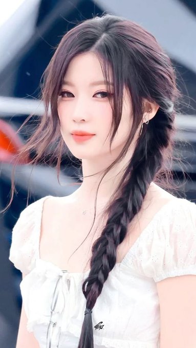 A portrait of a young woman with dark hair styled in a long side braid, wearing a white floral top, looking softly at the camera.