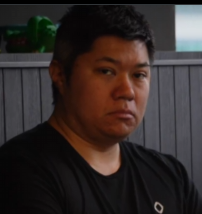A man with short dark hair in a black t-shirt looking at the camera with a serious, somewhat displeased expression in what appears to be a video or interview setting.
