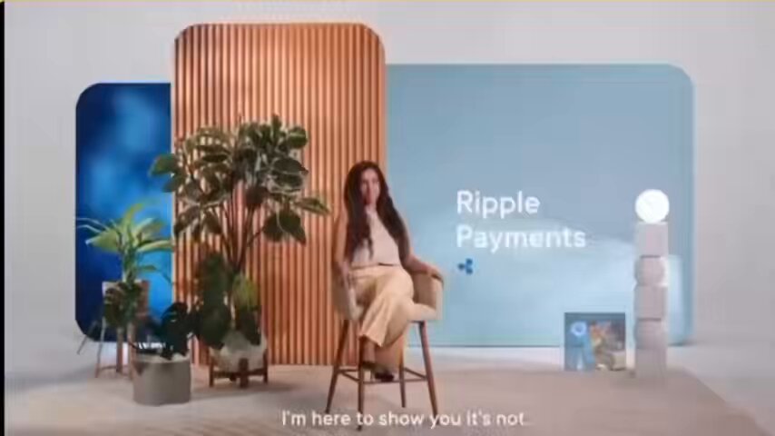 The image features a woman sitting on a chair with her legs crossed, wearing a white shirt. She appears to be talking about Ripple payments or discussing the global financial system. The background of the image includes a blue wall with a sign that says "RIPPLE" and "PAYMENTS", as well as some potted plants placed around the room. There is also an additional plant in the foreground, adding greenery to the scene.