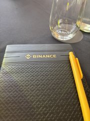A close-up of a dark notebook with a gold "BINANCE" logo, accompanied by a yellow pen and two glasses on a dark wooden surface.