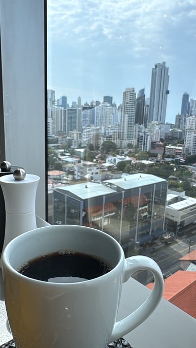 A white mug of black coffee sits on a windowsill, with a panoramic view of Panama City's modern skyline and skyscrapers visible through the glass, perfectly capturing the essence of a morning coffee overlooking the urban landscape.