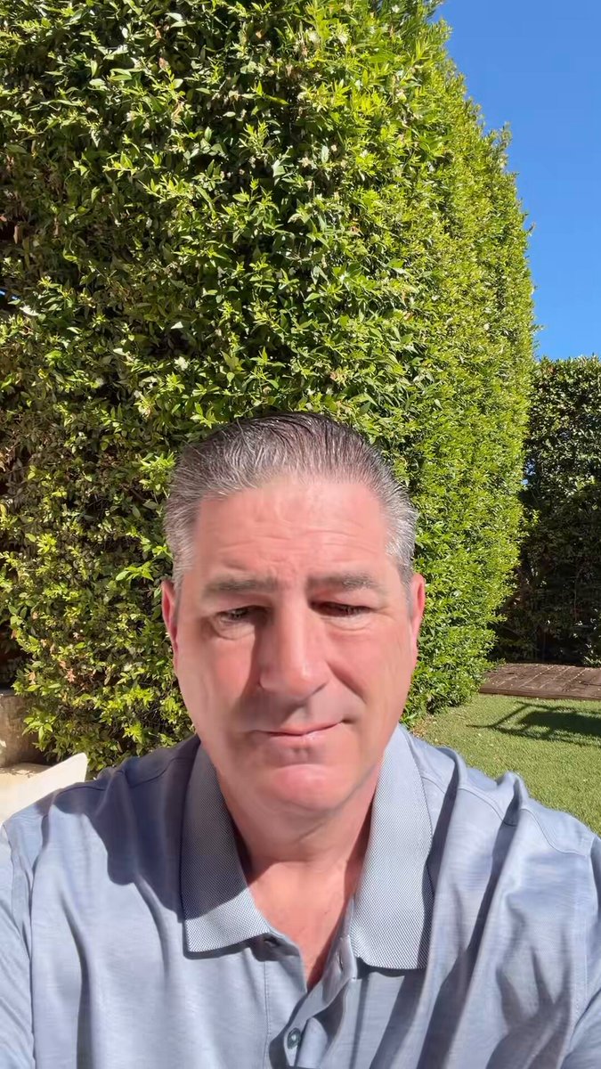 In this image, there's an older man sitting outside near some bushes with his head turned towards the camera, looking at us. He seems to be dressed formally, wearing a grey polo shirt. The photo has been edited to include text around the edges that says "Day 300". There is no visible action taking place in the scene, and it appears to be a calm and peaceful setting, with the man as the main subject of the photograph.