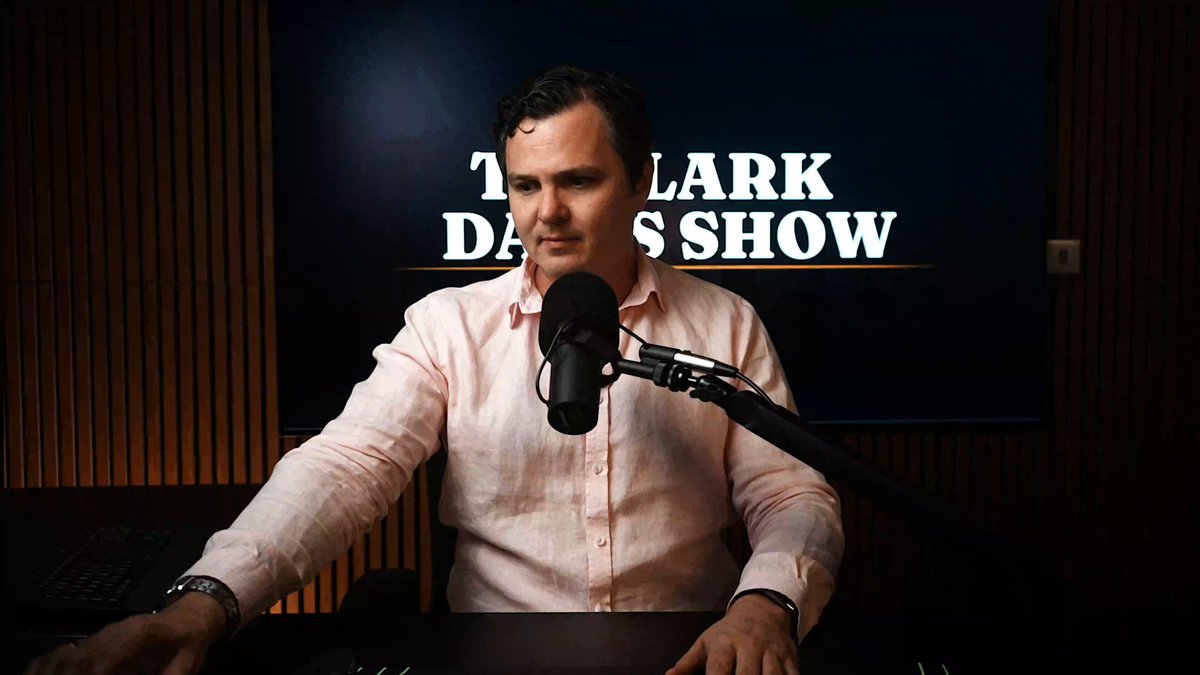 The image features a man sitting at a desk with a microphone, likely preparing for an interview or speaking event. The background shows a TV screen displaying "The Clark D. Show" logo, indicating that this might be a podcast or radio show being broadcasted on television. The man is wearing a pink shirt and appears to be focused on the task at hand.