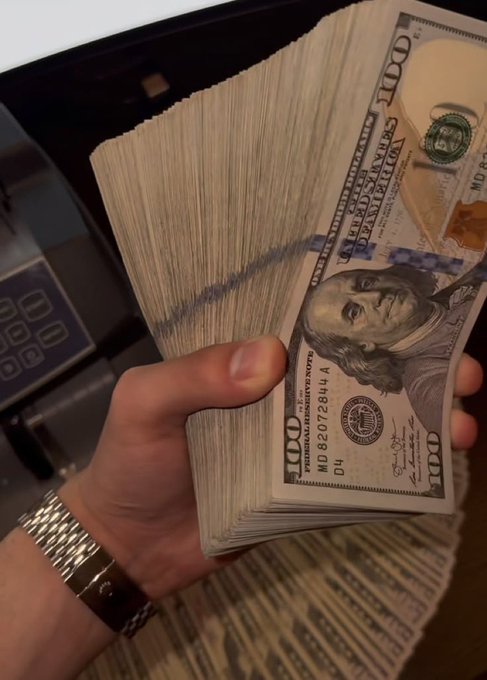 A close-up photo of a hand wearing a silver watch holding a thick stack of US $100 bills, with a money-counting machine visible in the background.