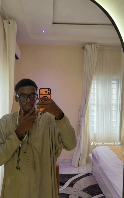 A young man with glasses, wearing a beige robe and chain necklace, takes a mirror selfie in a softly lit bedroom with white curtains, while the watermark "@nasthemessiah" displayed on the photo is being cited as evidence to expose the alleged scammer known as "Nasir" in a $17k+ and $5k+ online fraud scandal.