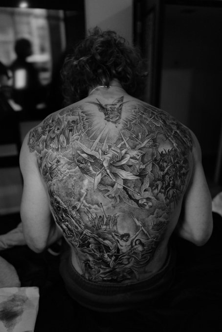 A man displays a massive, intricate black-and-gray tattoo covering his entire back, depicting a dramatic scene of angels, demons, and figures in a hellish landscape with a demonic figure looming at the top—visually capturing the infernal chaos of Dante's vision. The towering composition of writhing souls, stormy skies, and celestial warriors mirrors the post's description of being dragged through "the 7th layer of hell," embodying both the artistic tribute and the brutal journey that inspired it.