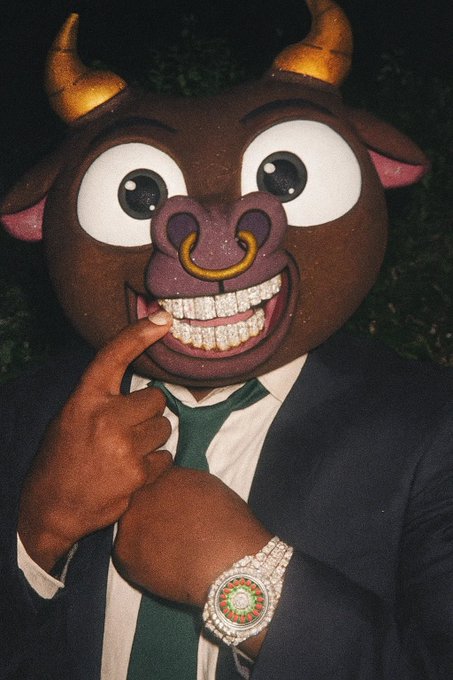A person wearing a large cartoon bull mascot mask with diamond-encrusted grillz, a green tie, and a luxurious Rolex-style watch, posing with a finger to the mask.