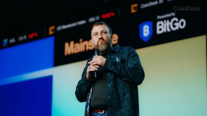 Cardano's Charles Hoskinson's address is shown giving a talk at a CoinDesk event sponsored by BitGo, as he reveals he is personally developing a new programming language for the blockchain. The backdrop displays "CoinDesk," "BitGo," and "Mainst" text during the blockchain-focused presentation.