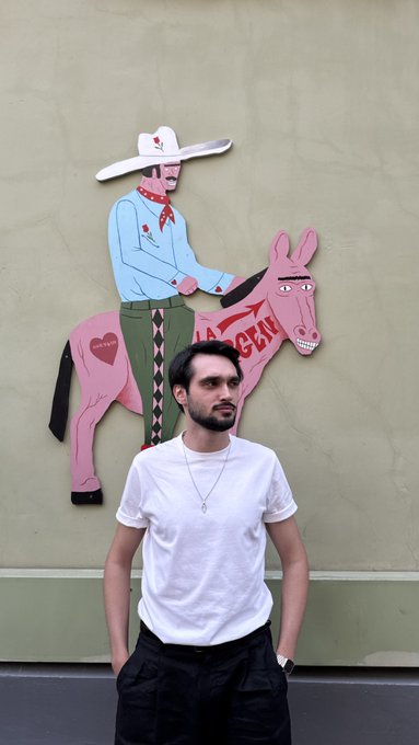 A young man in a white tee poses in front of a vibrant mural of a cowboy on a pink horse, matching the relaxed Sunday vibe of recharging—because nothing fixes a slow weekend quite like tacos. 🐴🌮
