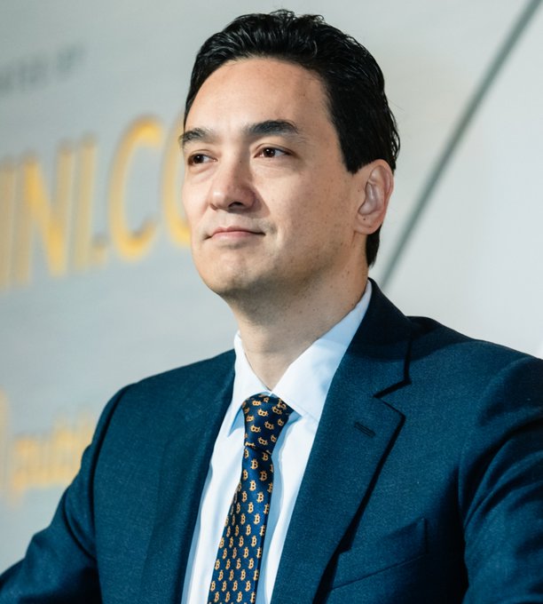 A portrait of a man in a dark blue suit and patterned tie standing in front of a blurred yellow sign.
