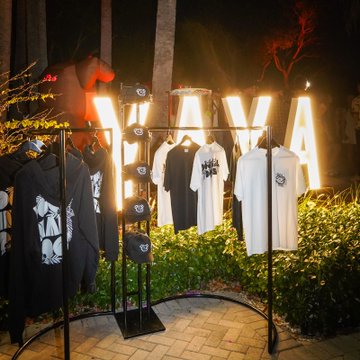 An outdoor nighttime display of clothing merchandise (t-shirts and hoodies) on black metal racks, illuminated by large lit-up 'G FUEL' marquee letters, with a horse statue visible in the background.