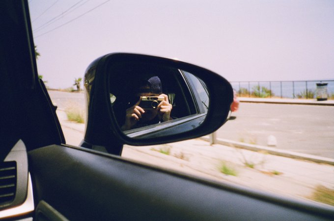 A mirror selfie taken through a car's side mirror, showing a person holding a camera with a blurred background of a road and fence.