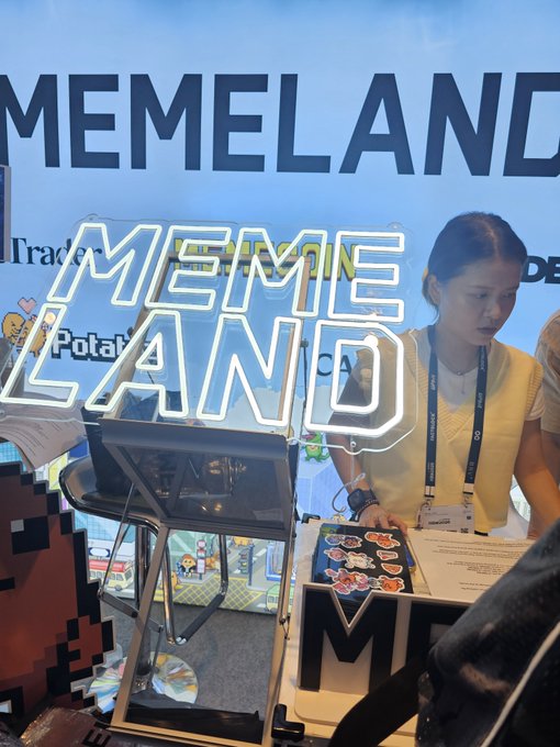 A woman stands behind a booth at what appears to be the "KNW" conference, with a large illuminated "MEMELAND" neon sign prominently displayed. She wears a lanyard and appears to be representing Memeland, the company behind the $MEME cryptocurrency mentioned in the caption, with promotional stickers and merchandise visible on the table.