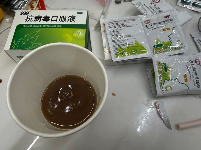 A paper cup contains a brown liquid next to Chinese medicine packaging, including a green box labeled "999 抗病毒口服液" (999 Antiviral Oral Liquid) and sachets of "双辛除湿败毒颗粒" (Shuang Xin Dampness-Removing Detoxifying Granules), illustrating the blogger's remedy that helped treat their illness.