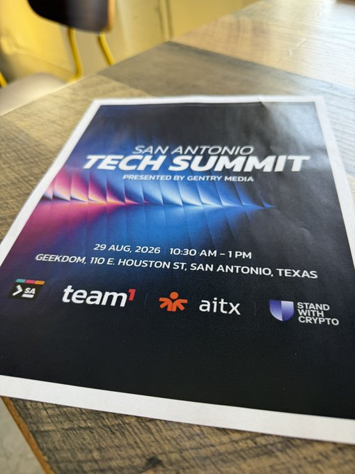 A flyer on a wooden table advertises the 'San Antonio Tech Summit' presented by Gentry Media, taking place on 29 Aug. 2026, 10:30 AM - 1 PM at Geekdom, 110 E. Houston St., San Antonio, Texas, with sponsors team¹, aitx, and Stand With Crypto.