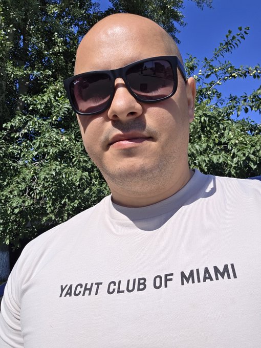 A bald man wearing dark sunglasses and a white t-shirt with 'YACHT CLUB OF MIAMI' text, standing outdoors against a blue sky with green foliage.