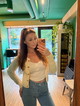 A young woman with long brown hair takes a mirror selfie wearing a cream-colored cardigan and blue jeans in a brightly lit room with green walls. The image appears to be a casual personal post unrelated to finance, showing her relaxed summer style.