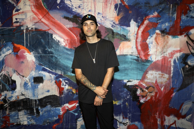 A young man wearing a black cap, black t-shirt, and silver necklace stands with crossed arms in front of a large abstract painting with red, blue, and white brushstrokes.