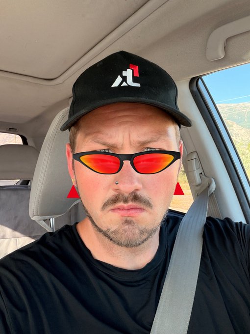 A selfie of a man in a car wearing a black cap, red-tinted sunglasses, and digital red triangle edits on his cheeks and earrings.