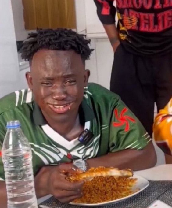 A young man with dreadlocks, wearing a green and white jersey, sits at a table crying while holding a piece of food over a plate of reddish rice, with a water bottle beside him.
