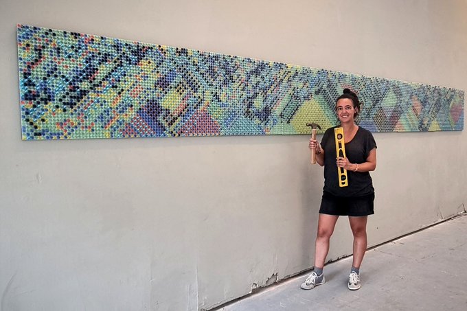 A smiling woman in a black dress stands next to a large colorful wall installation made of circular bottle caps, holding a yellow level and a hammer.