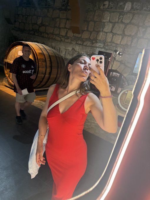 A mirror selfie of a woman in a red dress holding a smartphone with a heart emoji on the case, while a man wearing a red and black patterned shirt stands in the background near wooden barrels in what appears to be a wine cellar.