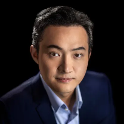 A professional headshot of a man with dark hair wearing a navy blazer and light blue shirt against a black background.