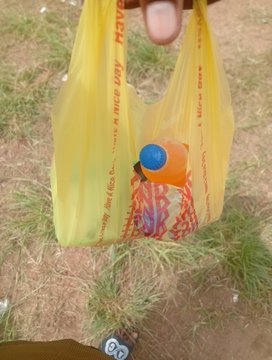 A hand holds up a yellow plastic shopping bag containing what appears to be food items, including a bottle with an orange cap, set against a grassy outdoor background—likely a campsite. The image aligns with the post's mention of being "in camp" and proudly showing off a sponsor ("web3warrior"), suggesting the bag may contain sponsored refreshments enjoyed during the outdoor outing.