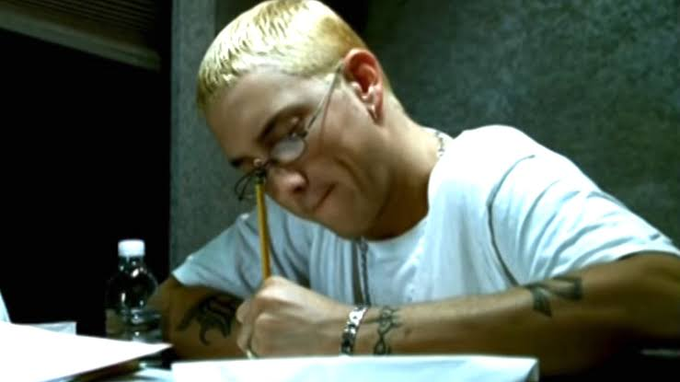 A man with short blonde hair and glasses, wearing a white t-shirt and a chain necklace, writing with a pencil at a desk with a water bottle nearby.