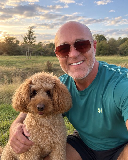 A bald man wearing aviator sunglasses and a teal Under Armour shirt smiles warmly while posing with a curly-haired brown poodle in a grassy field at sunset.