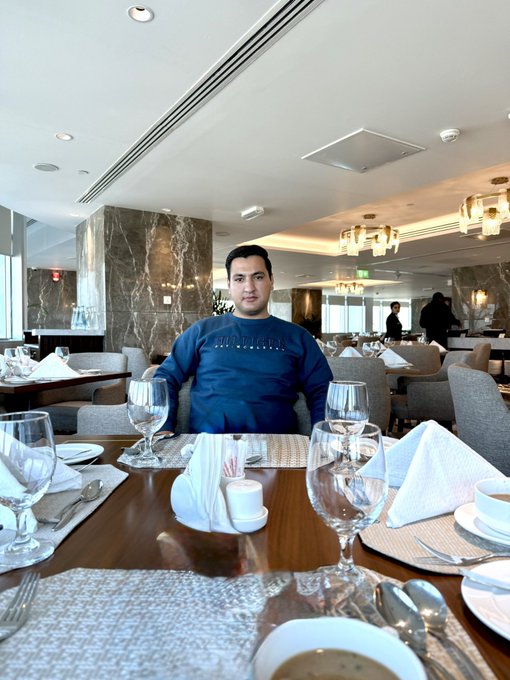 A man wearing a navy Tommy Hilfiger sweatshirt sits at an elegantly set dining table in an upscale restaurant, smiling at the camera with the cheerful caption "GOOD MORNING EVERYONE. Are you ready to win today? Who will say 'GM back'?" encouraging his audience to greet him back in the comments.