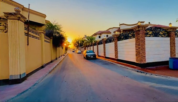 A residential street at sunset with luxury gated villas on both sides, the warm golden light enhancing the aspirational atmosphere. The image visually echoes the post's theme of ambition and reward, symbolizing the upscale lifestyle the author is striving toward as they pursue the Top 300 rewards on NomismaNetwork.