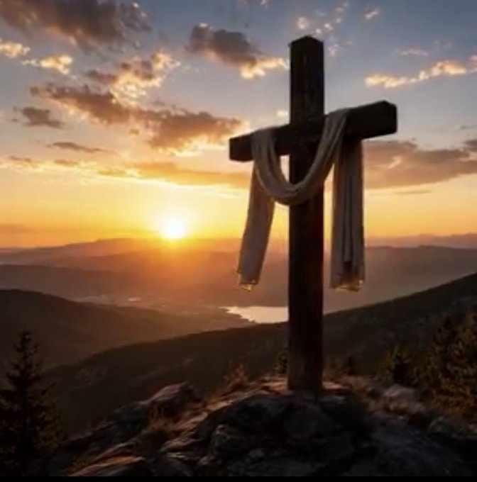 A wooden cross draped with a white cloth stands on a rocky outcrop overlooking a valley at sunset, with a lake and mountains in the distance.
