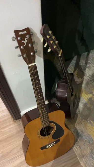 Two Yamaha acoustic guitars leaning against a wall on a wooden floor, with a curtain visible in the background.