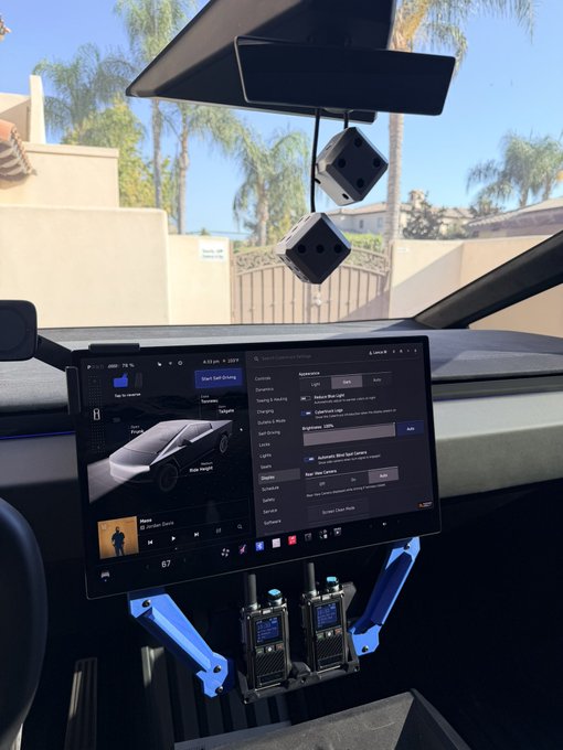 Interior view of a vehicle featuring a large central touchscreen displaying vehicle settings (Suspension: Light, Lights: Ambient Blue (Low), etc.) and media controls, with fuzzy dice hanging from the rearview mirror and palm trees visible outside.