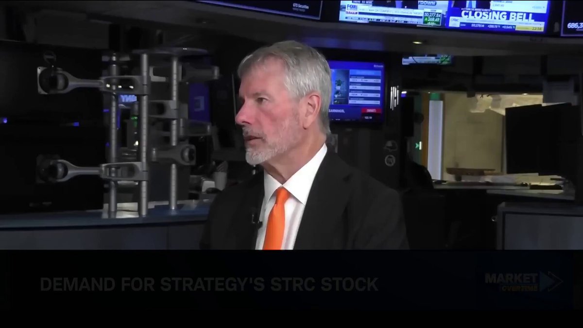 The image shows a man wearing a suit and tie standing behind a desk with a computer monitor in front of him. The background features a TV screen displaying stock market information along with a news bulletin about closing bell times for stocks. The text on the image reads "Demand for strategy's strc stock".