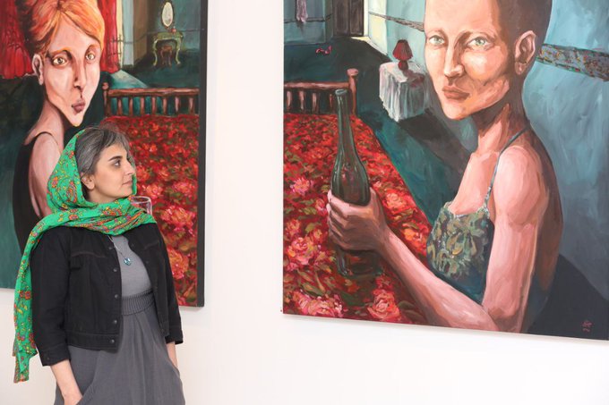 A woman wearing a green headscarf and dark jacket stands in an art gallery between two large paintings depicting women in interior settings.